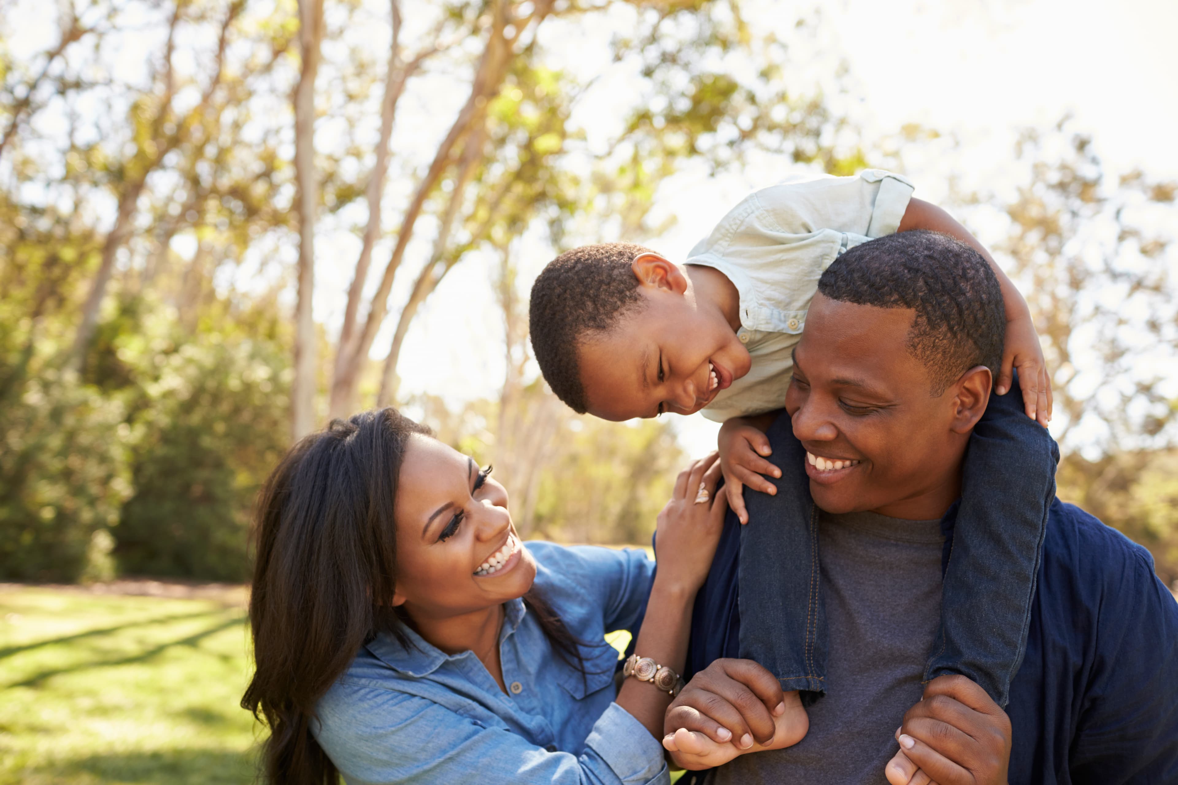 Two adults laughing outdoors with a young child riding on one adult's shoulders